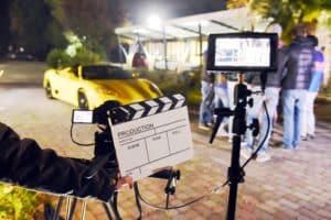Operator holding clapperboard during the production of short film outdoor in the night with sportive yellow car and actor on stage. Focus on the clapperboard and monitors